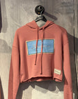 Pink hoodie with a blue logo on a hanger against a textured wall.
