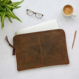 Brown leather laptop pouch on a light surface with a plant, glasses, and cup in the background