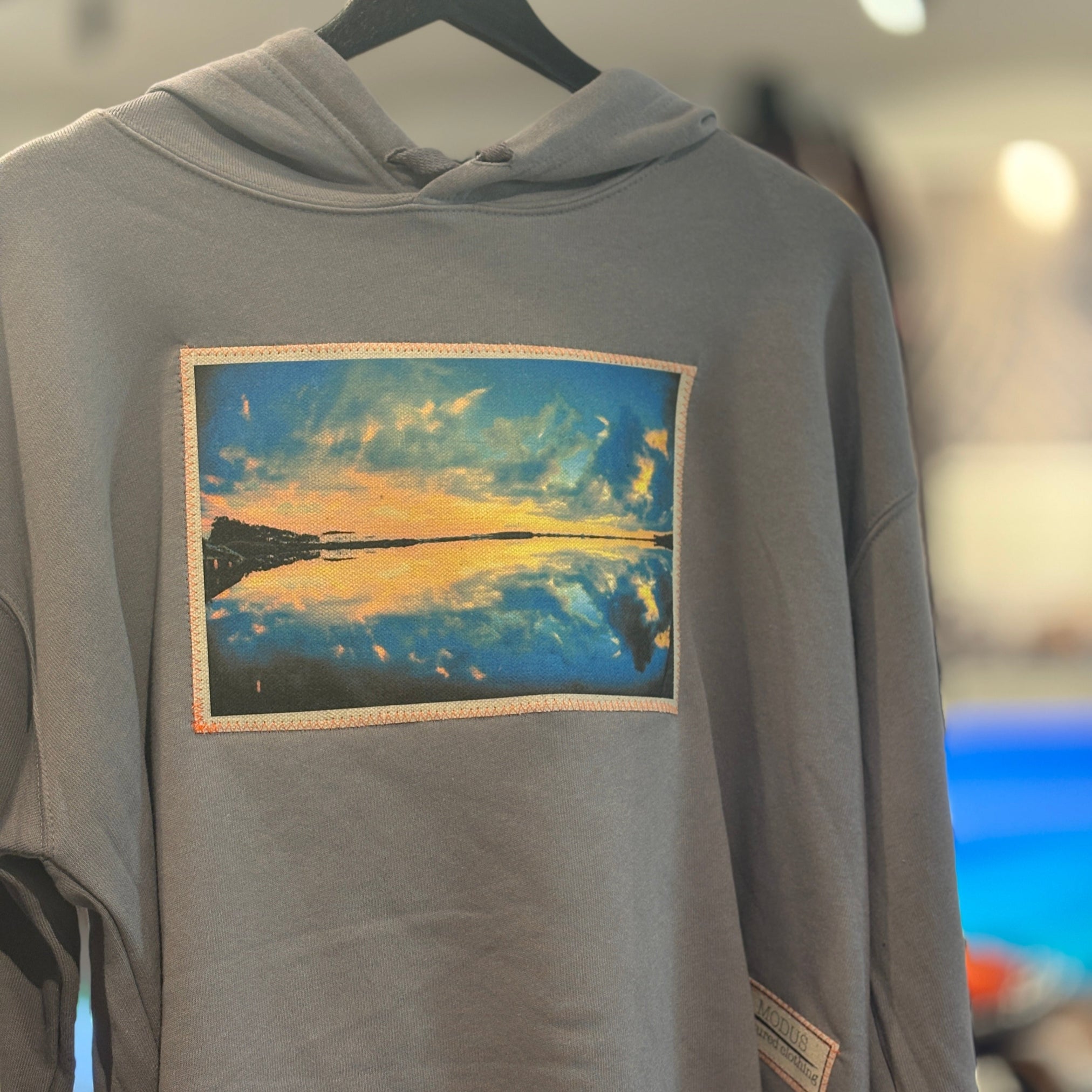 Gray hoodie with a graphic design of a sunset and clouds on a hanger.