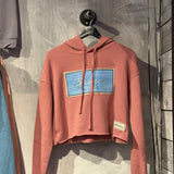 Pink hoodie with a blue logo on a hanger against a textured wall.
