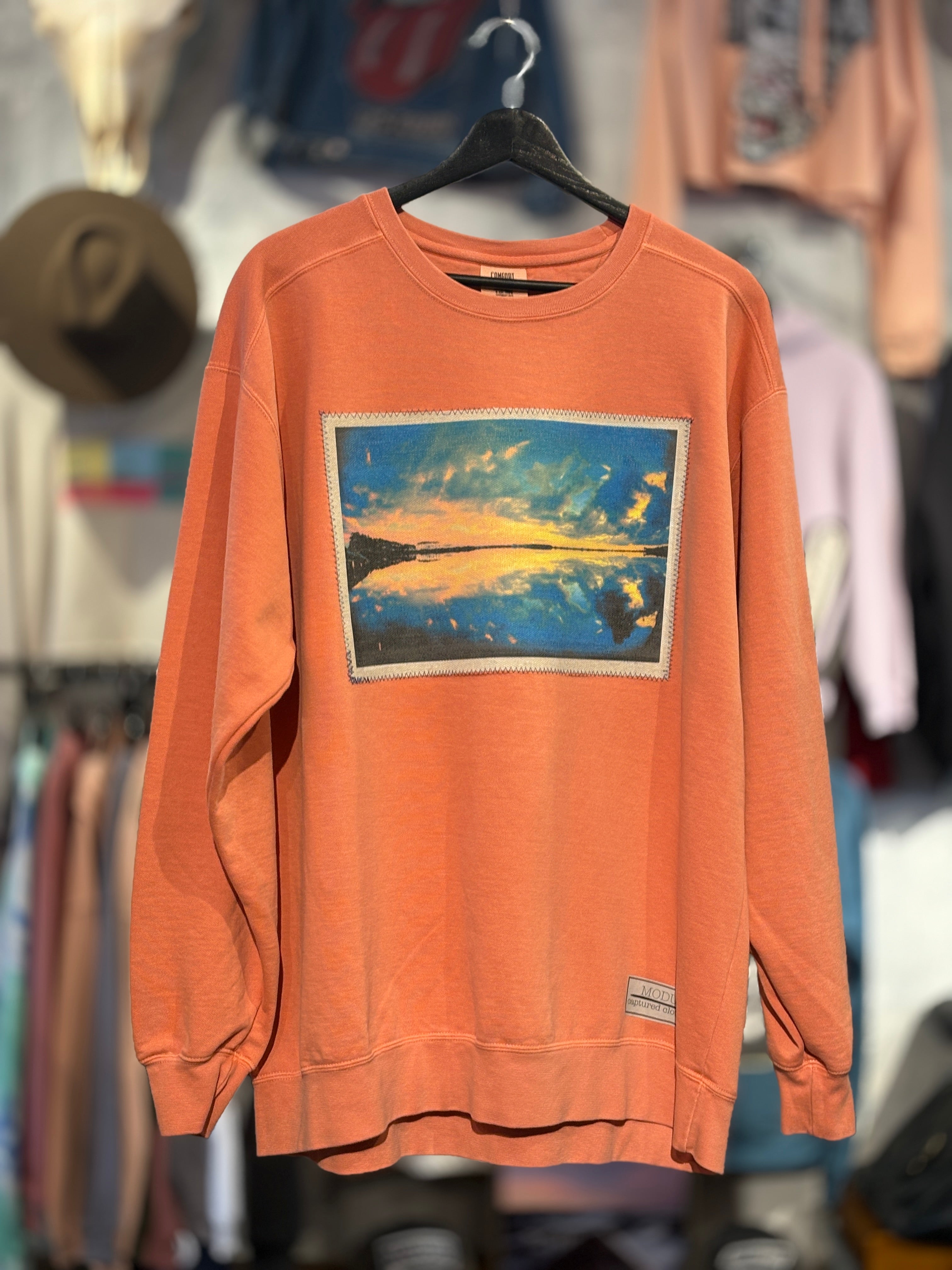 Orange sweatshirt with a graphic design of a sunset over water, hanging on a rack with other clothing in the background.