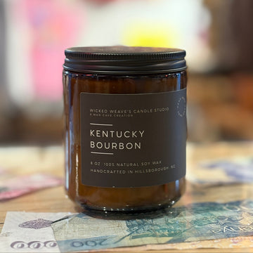 Candle labeled 'Kentucky Bourbon' from Wicked Weaver Candle Studio on a table with a blurred background.