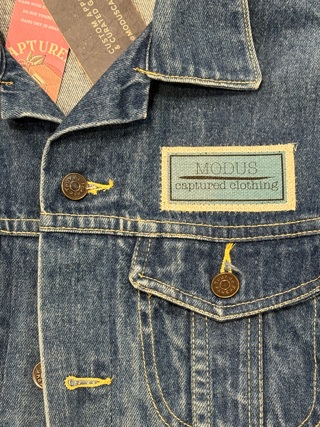 Close-up of a denim jacket with 'Modus Captured Clothing' branding and visible buttons.