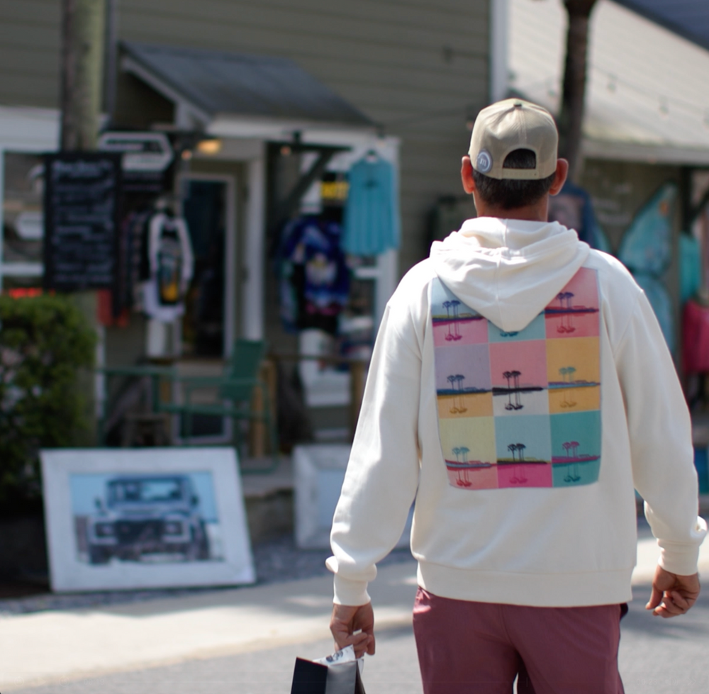 Person wearing a white hoodie with colorful palm tree pattern walking on a street in front of captured clothing the store. 