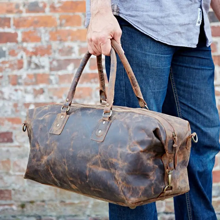 Person holding a brown leather duffel bag against a brick wall.