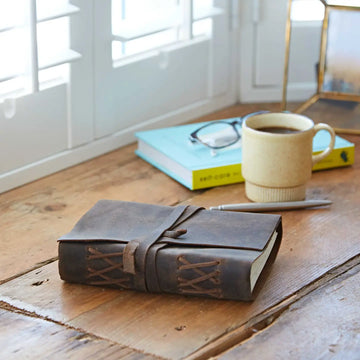 Brown leather-bound notebook on a wooden surface with a cup of coffee and glasses in the background.