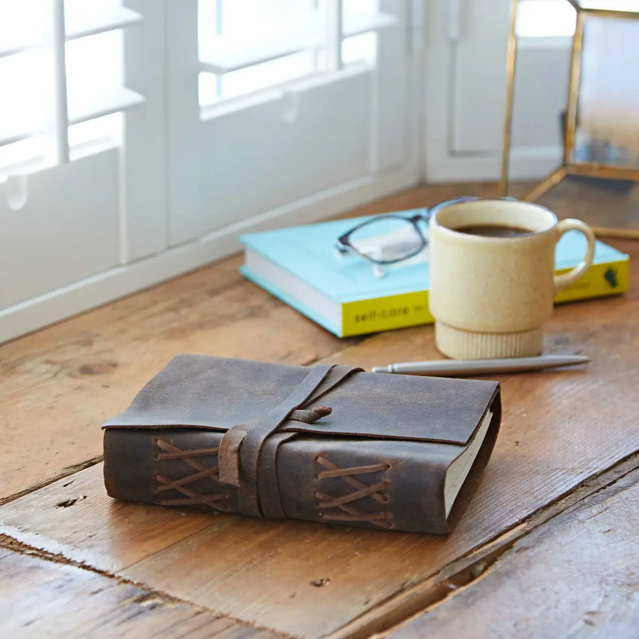 Brown leather-bound notebook on a wooden surface with a cup of coffee and glasses in the background.