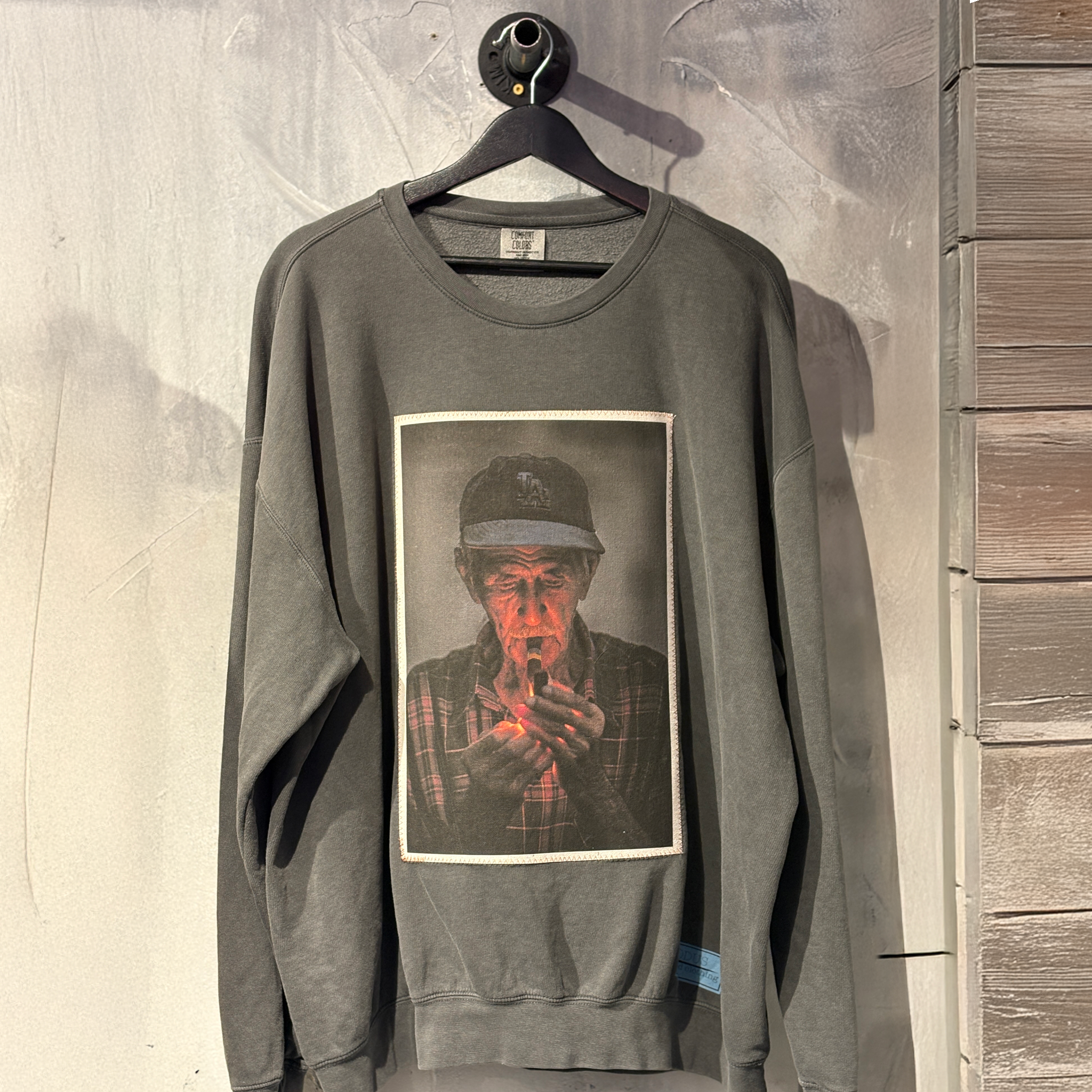 Gray sweatshirt with a graphic print of a person smoking a cigarette, hanging on a wooden surface.