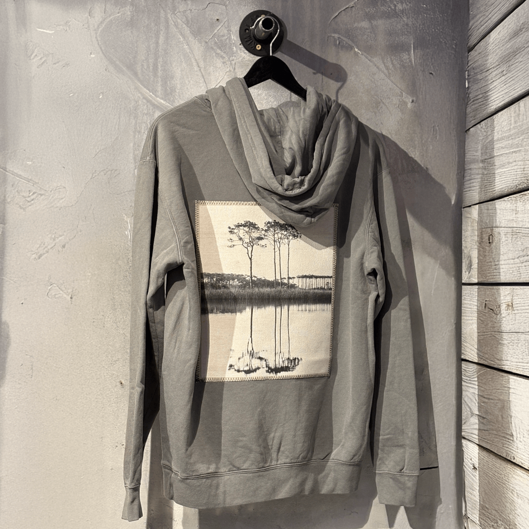Gray hoodie with a western lake graphic on a hanger against a textured wall.