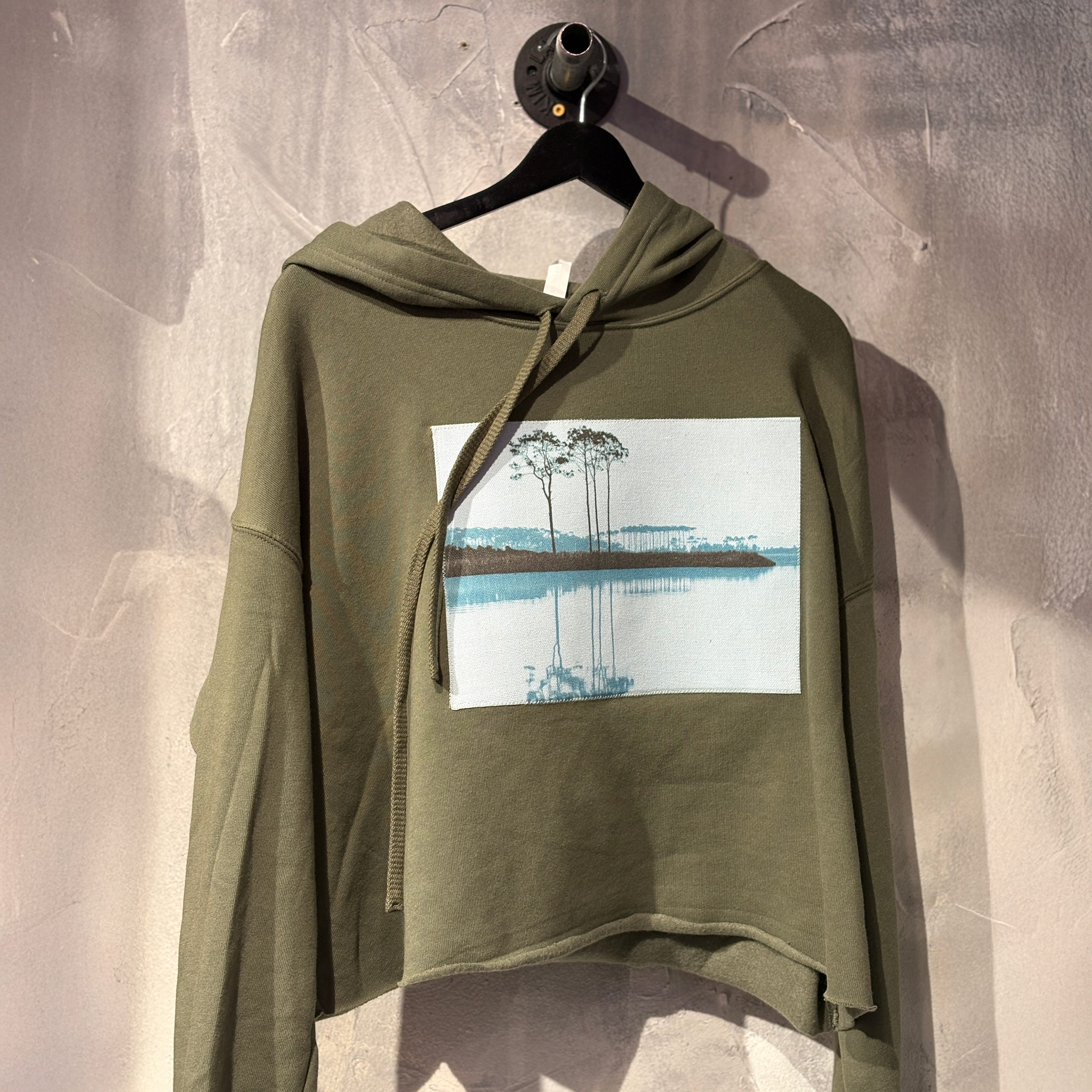 Green hoodie with a printed design of a lake on a hanger against a textured wall.