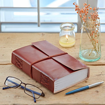 Brown leather-bound notebook on a wooden surface with glasses and a pen nearby